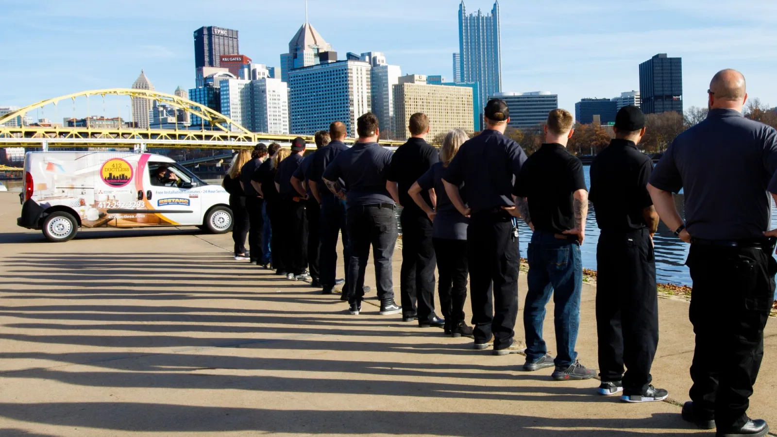 Group of people standing in line by riverfront with city skyline and bridge in background on sunny day