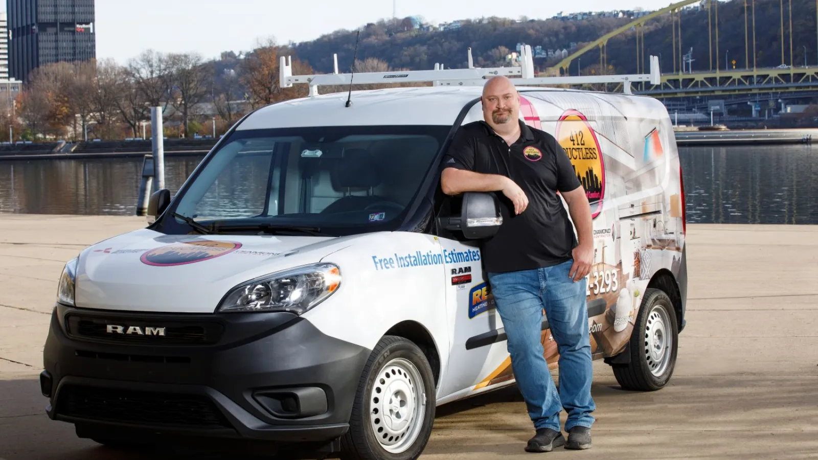 Man in black shirt stands by branded RAM van offering free installation estimates by a riverside with city skyline background
