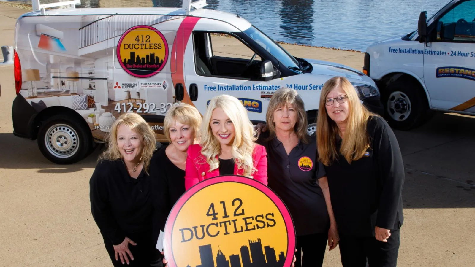 Five women posing with a 412 Ductless sign near company vans by a riverside urban skyline