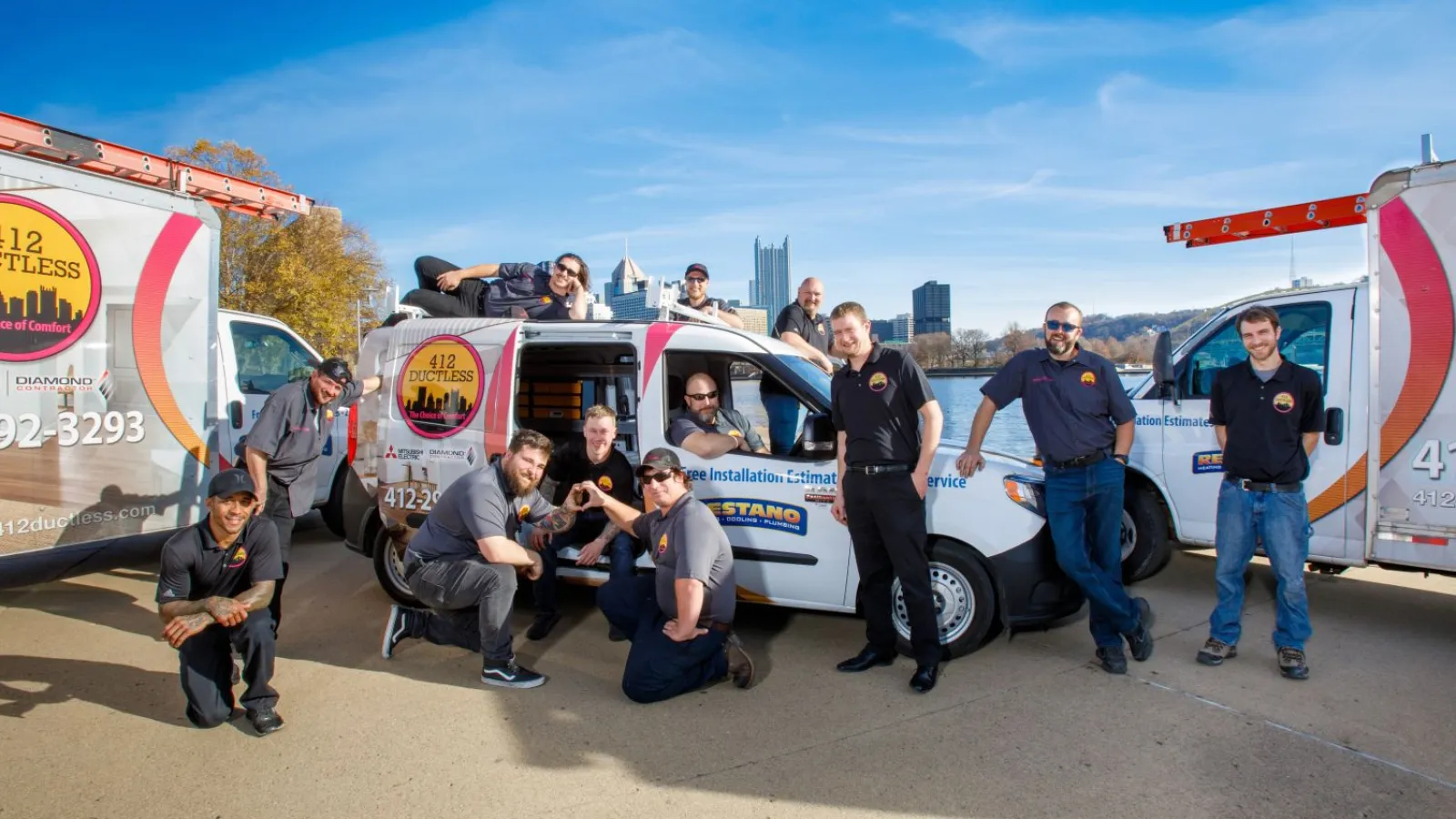Team of HVAC professionals posing with 412 Ductless branded service vans under clear blue sky in urban setting.