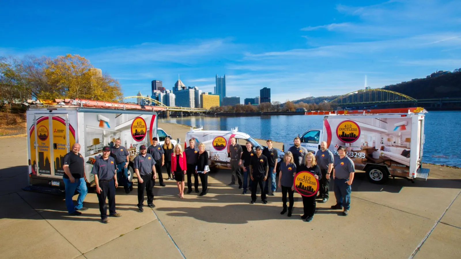 Team of 412 Auction staff posing outdoors with branded trucks by riverside and city skyline in background