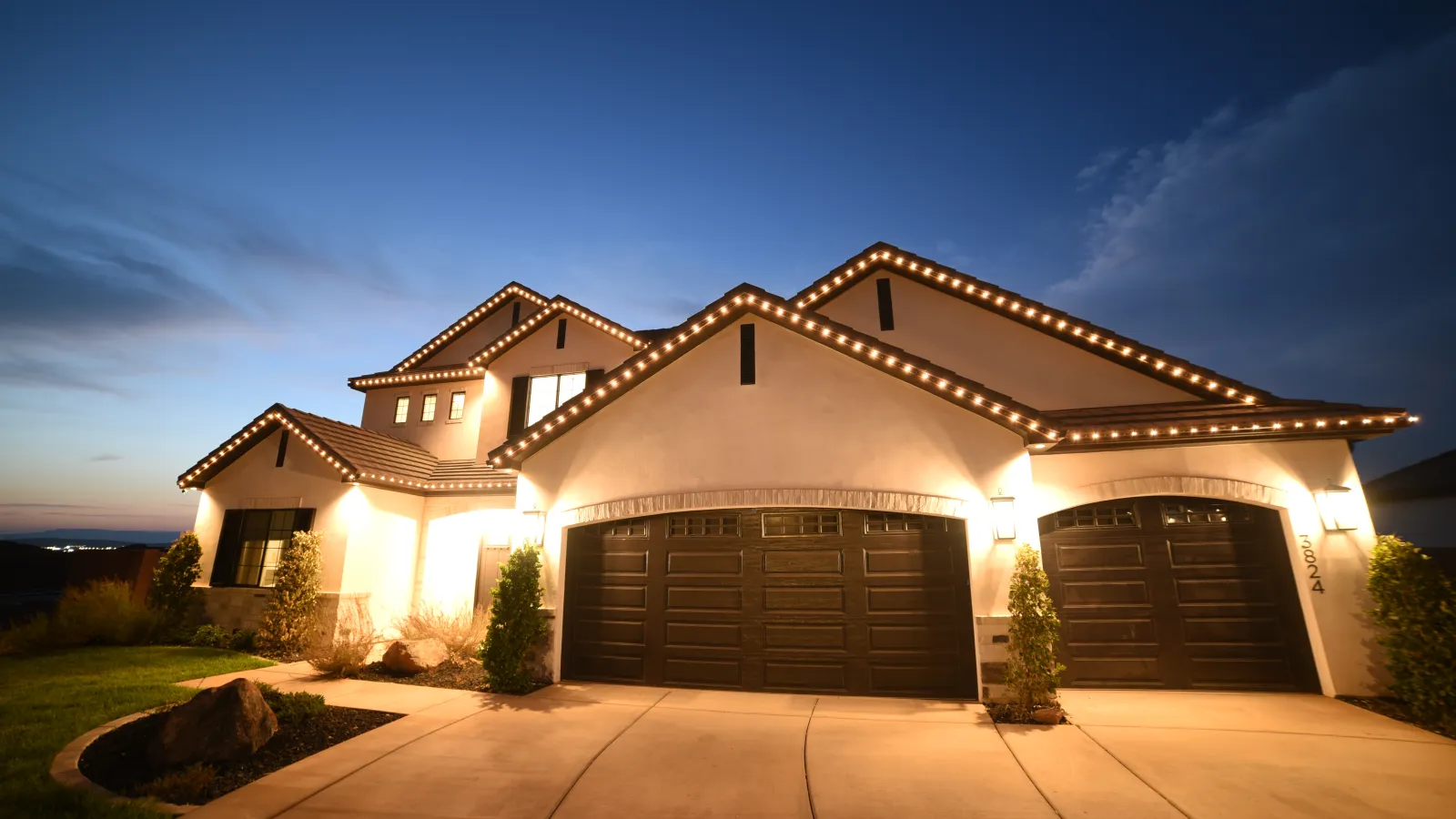 Modern two-story home at dusk with warm exterior lights and decorated roofline lighting.