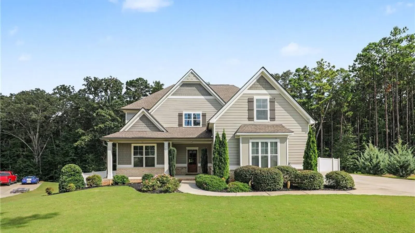 Two-story beige suburban house with a front porch, manicured lawn, and trees in the background under a blue sky