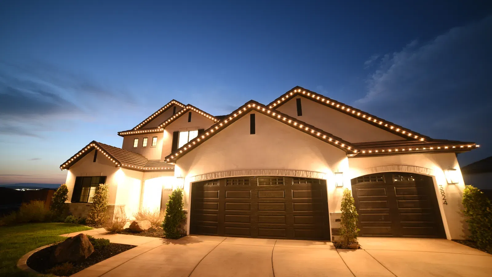 Modern suburban house with garage doors, exterior lights, and roofline decorated with string lights at dusk.