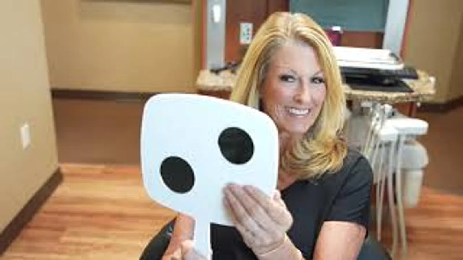 Smiling woman holding dental shade guide in a modern dental clinic with equipment in background