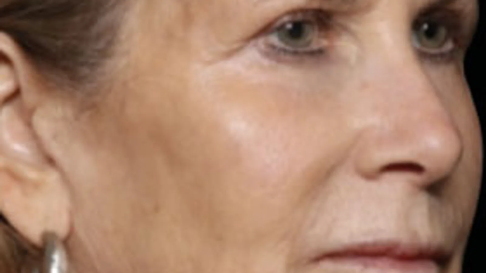 Close-up of an older woman’s face showing wrinkles and silver hoop earrings against a black background.