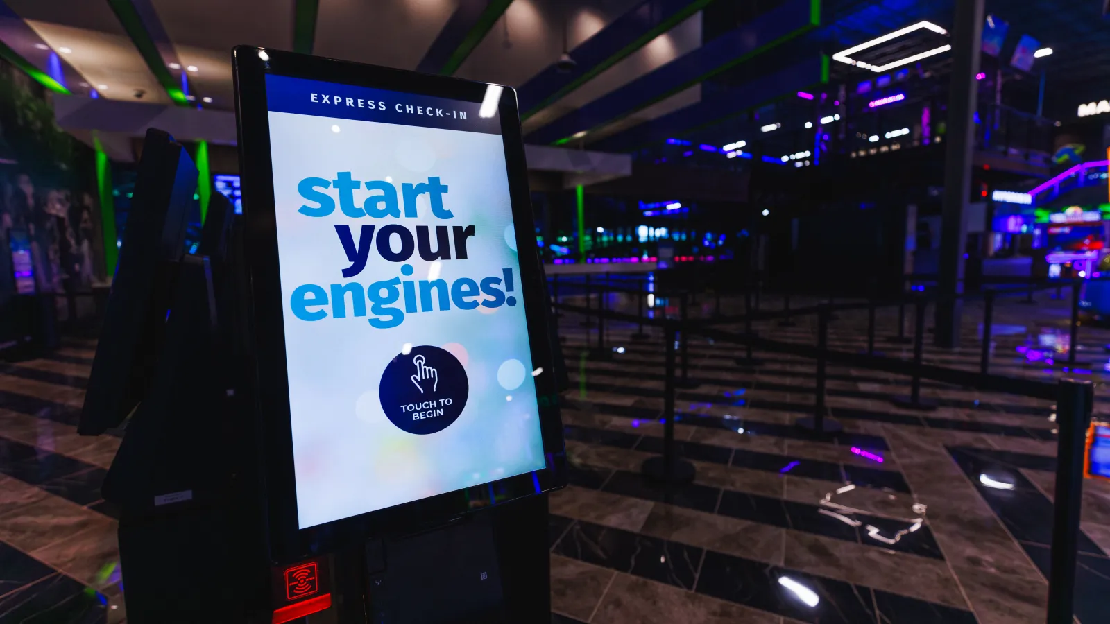 Touch screen kiosk in a dark entertainment center displaying start your engines for express check-in.