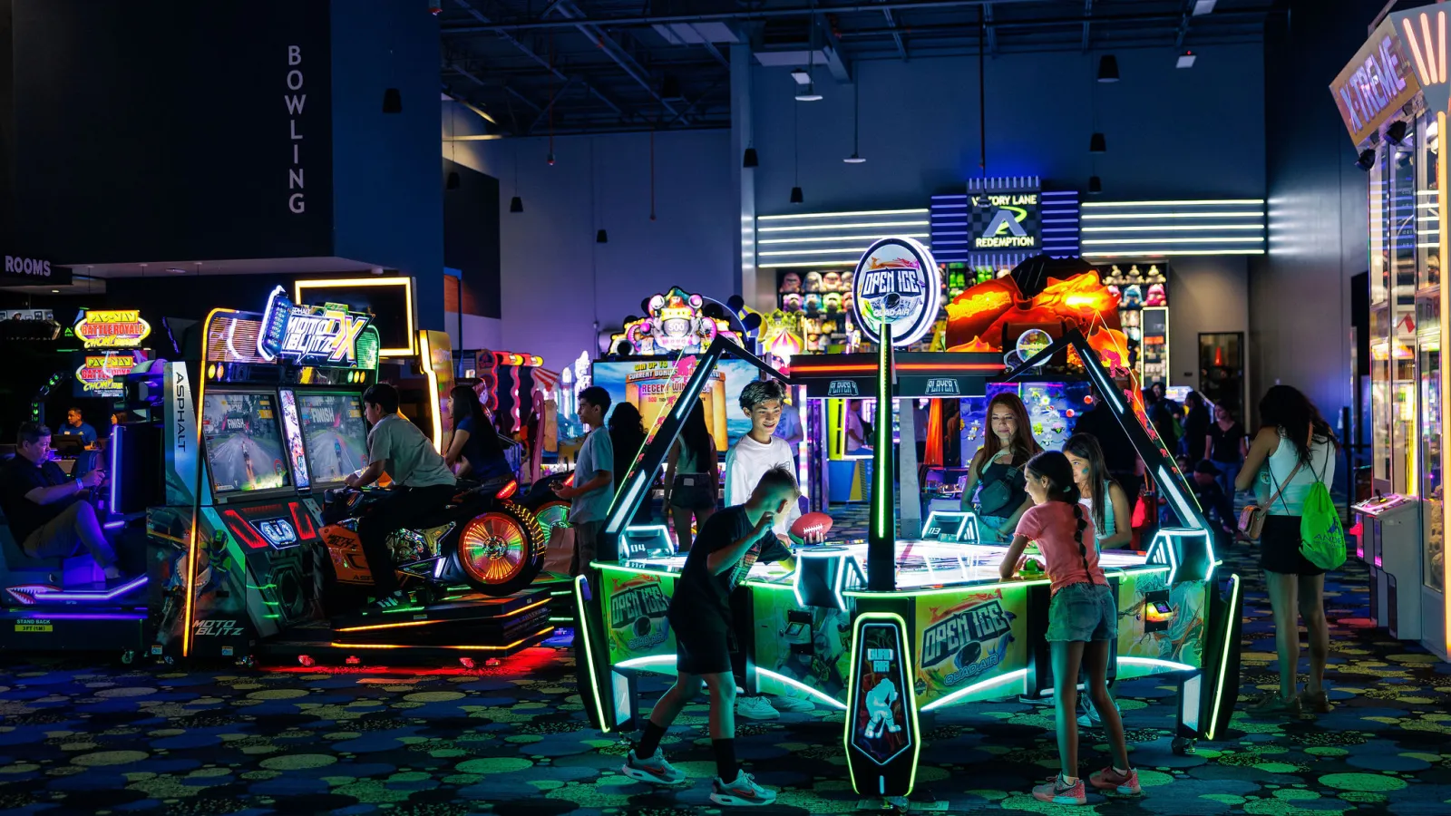 Kids and adults playing air hockey and arcade games in a neon-lit indoor entertainment arcade.