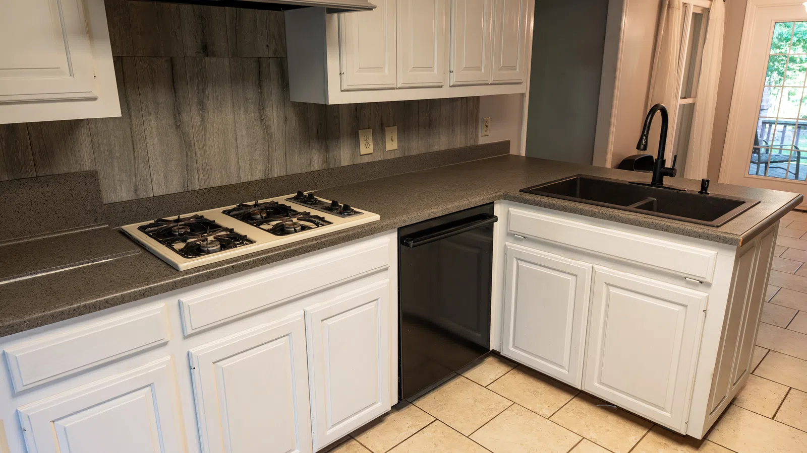 Modern kitchen with white cabinets, black countertop, gas stove, black sink, and tiled floor with back door view.