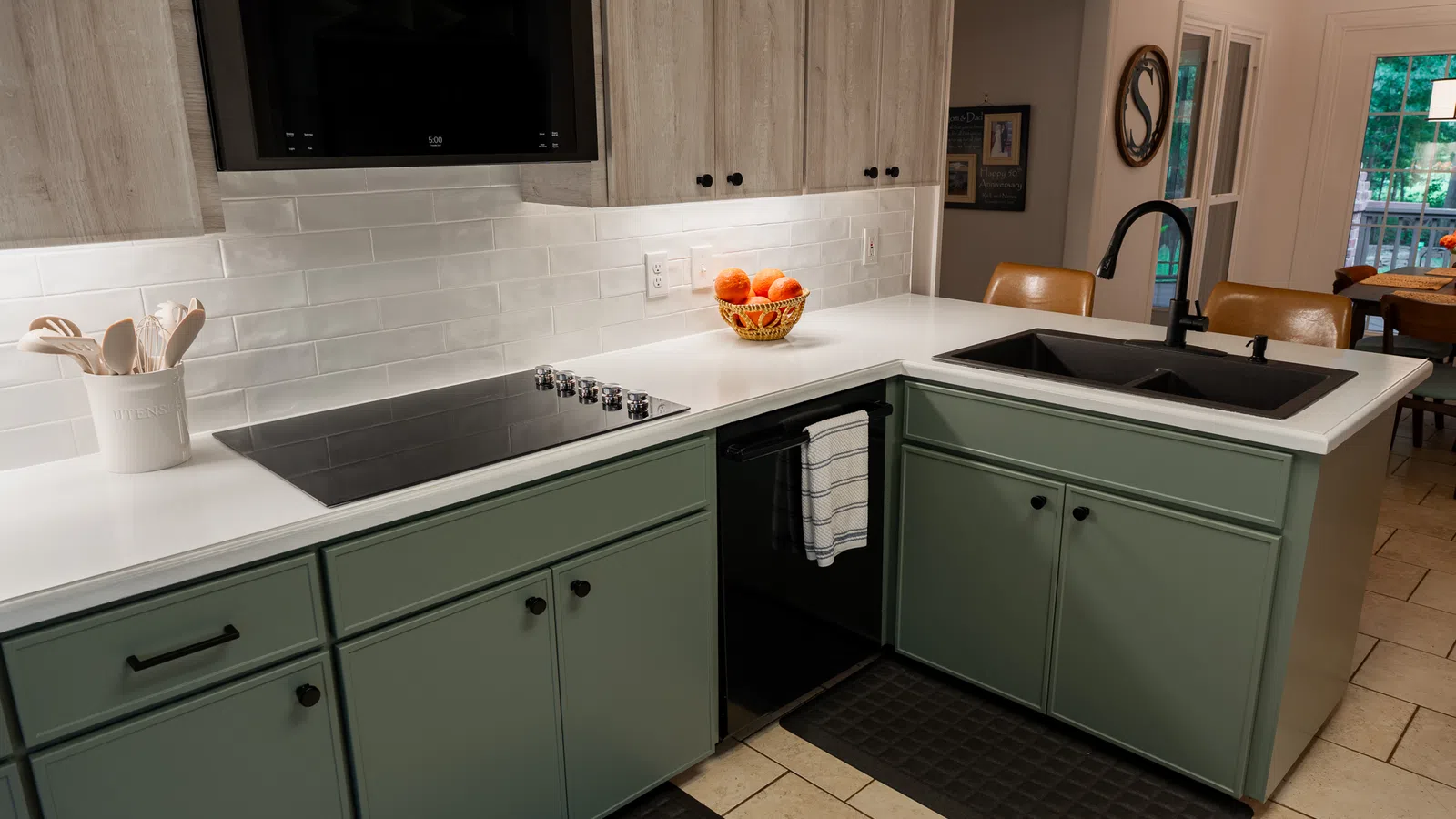 Modern kitchen with green lower cabinets, white countertops, black sink, stovetop, and a basket of oranges.