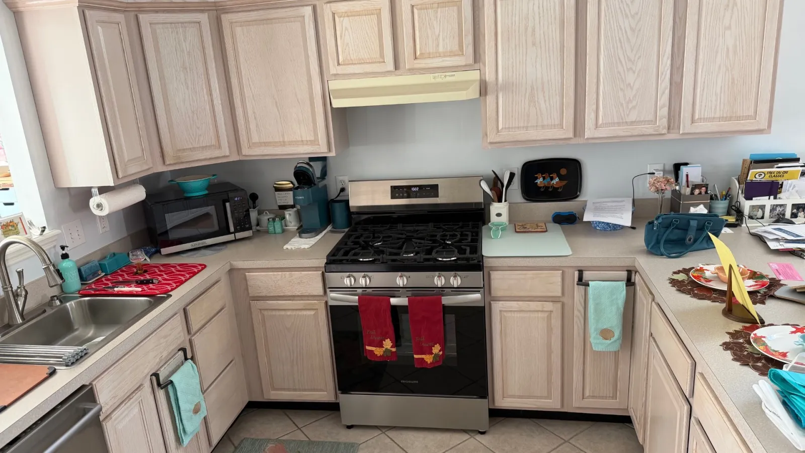 Light wood kitchen cabinets surround a stainless steel stove and sink in a bright, tidy kitchen with colorful towels.