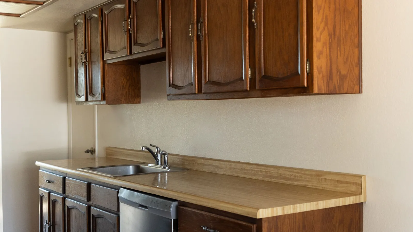 Kitchen countertop with wooden cabinets, stainless steel sink, and dishwasher in a simple beige room