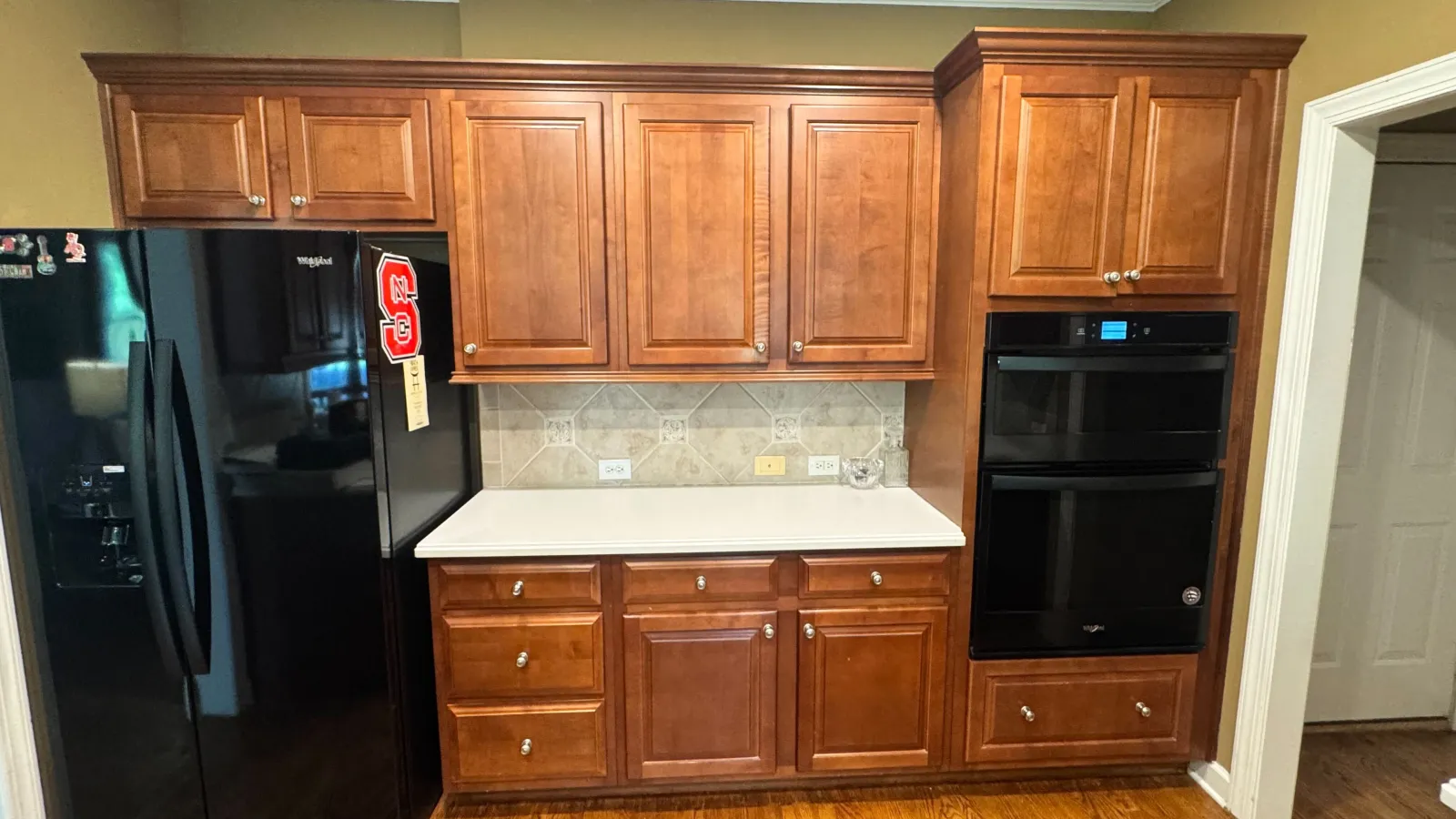 Kitchen wall with wooden cabinets, black refrigerator, built-in oven, white countertop, and hardwood floor.