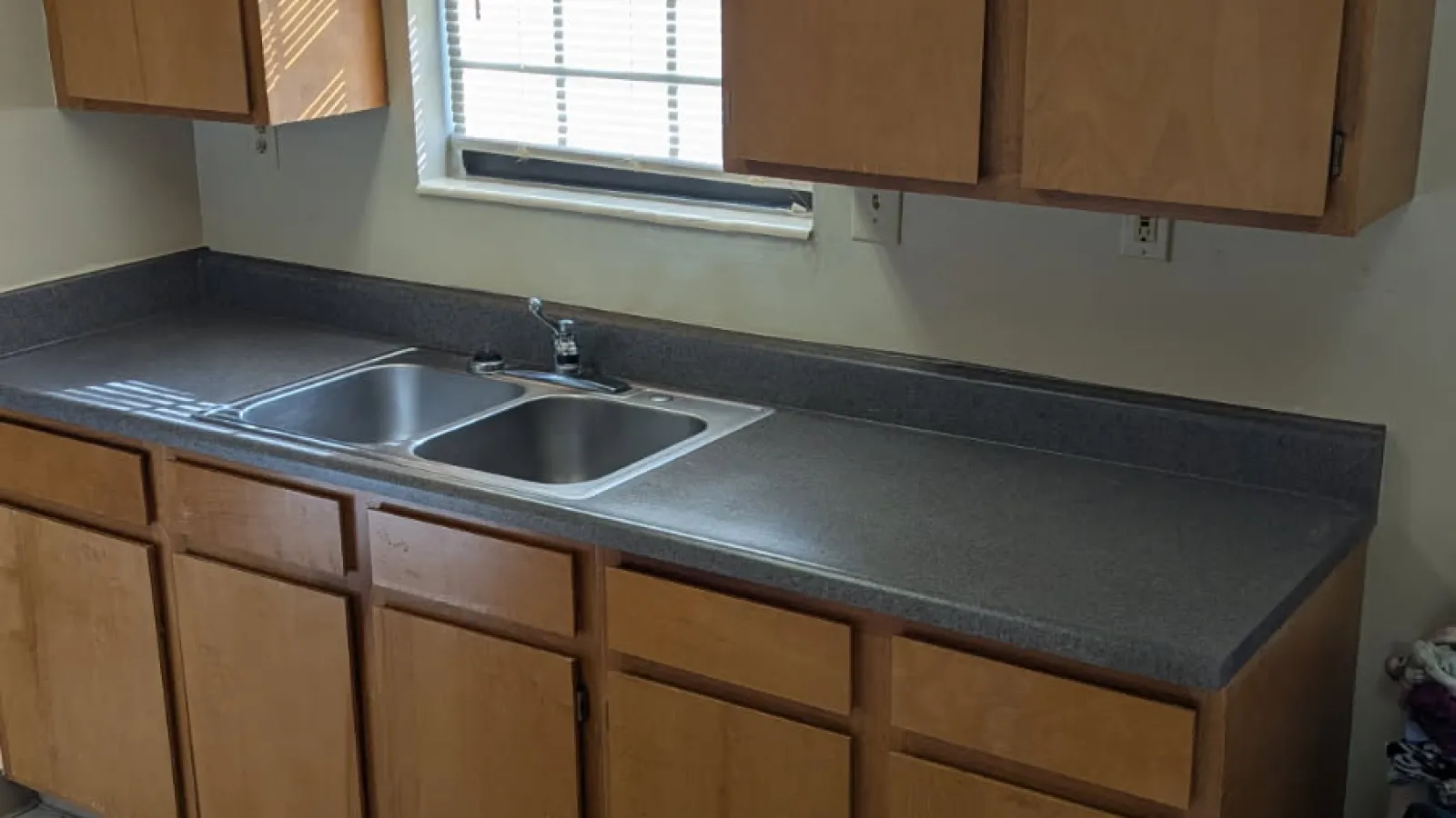 Kitchen with wooden cabinets, gray countertops, double sink below window with blinds, and vinyl flooring.