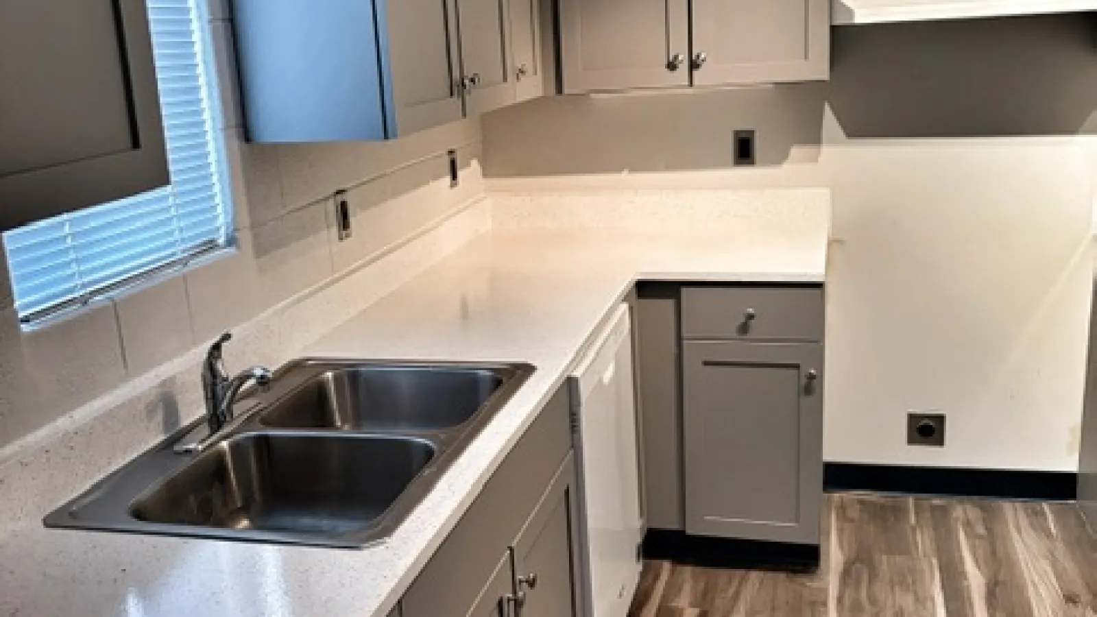 Modern kitchen with gray cabinets, white countertops, double stainless steel sink, and wood-look flooring.