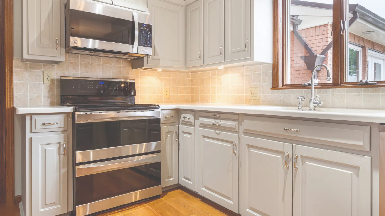 Bright kitchen with white cabinets, stainless steel oven and microwave, beige tiled backsplash, and wooden floor.