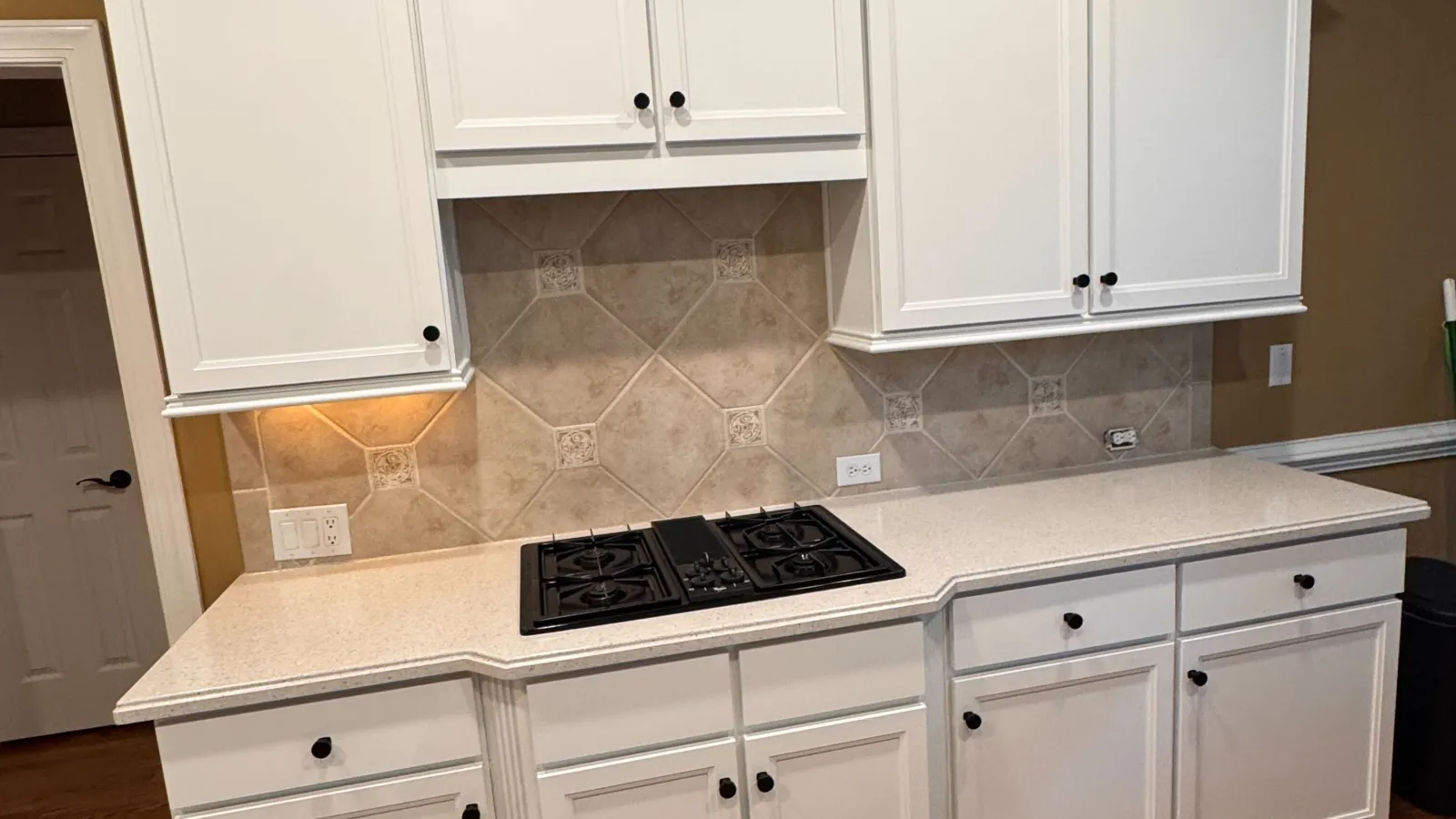 Modern kitchen with white cabinets, beige tiled backsplash, gas stovetop, and hardwood flooring with black floor mat.