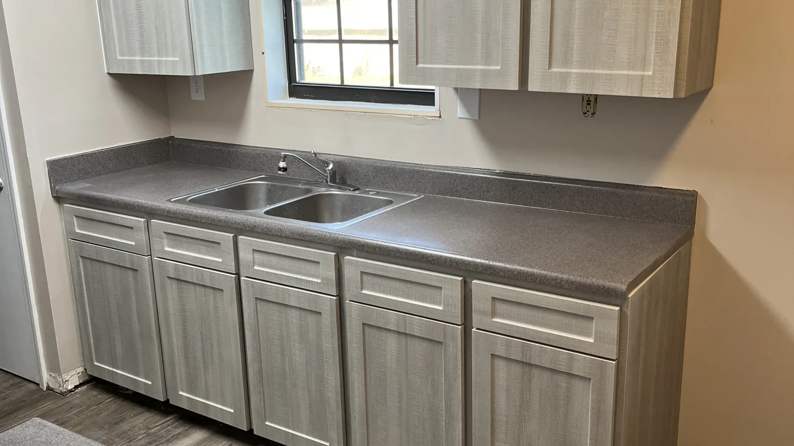Modern kitchen with light wood cabinets, gray countertops, stainless steel sink, and wood grain flooring.