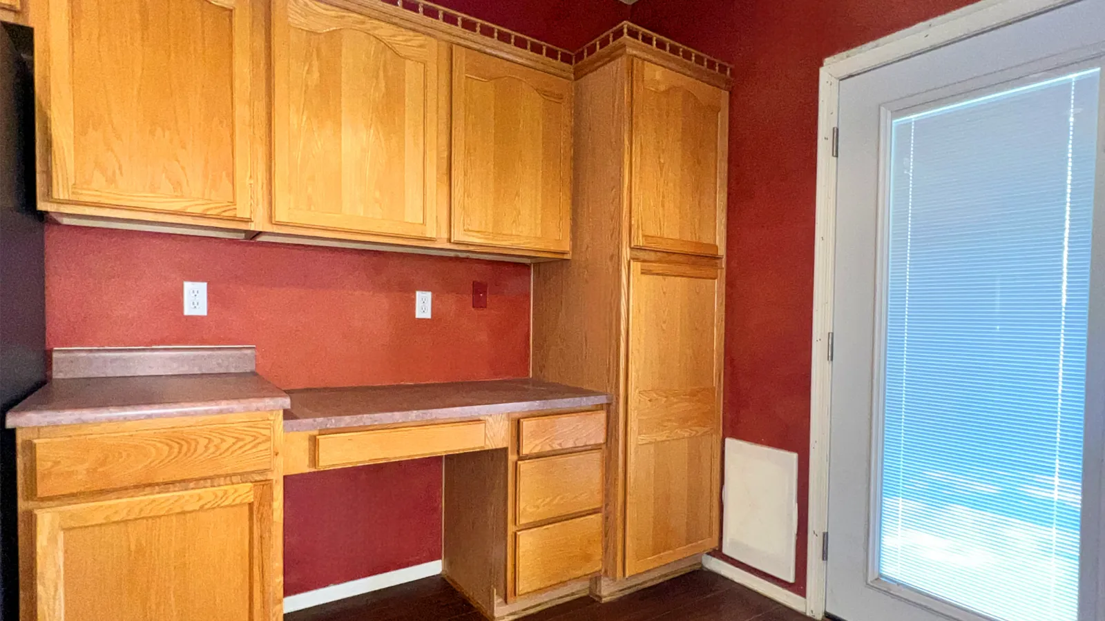 Kitchen corner with oak cabinets, dark wood floor, maroon walls, and white door with built-in blinds.