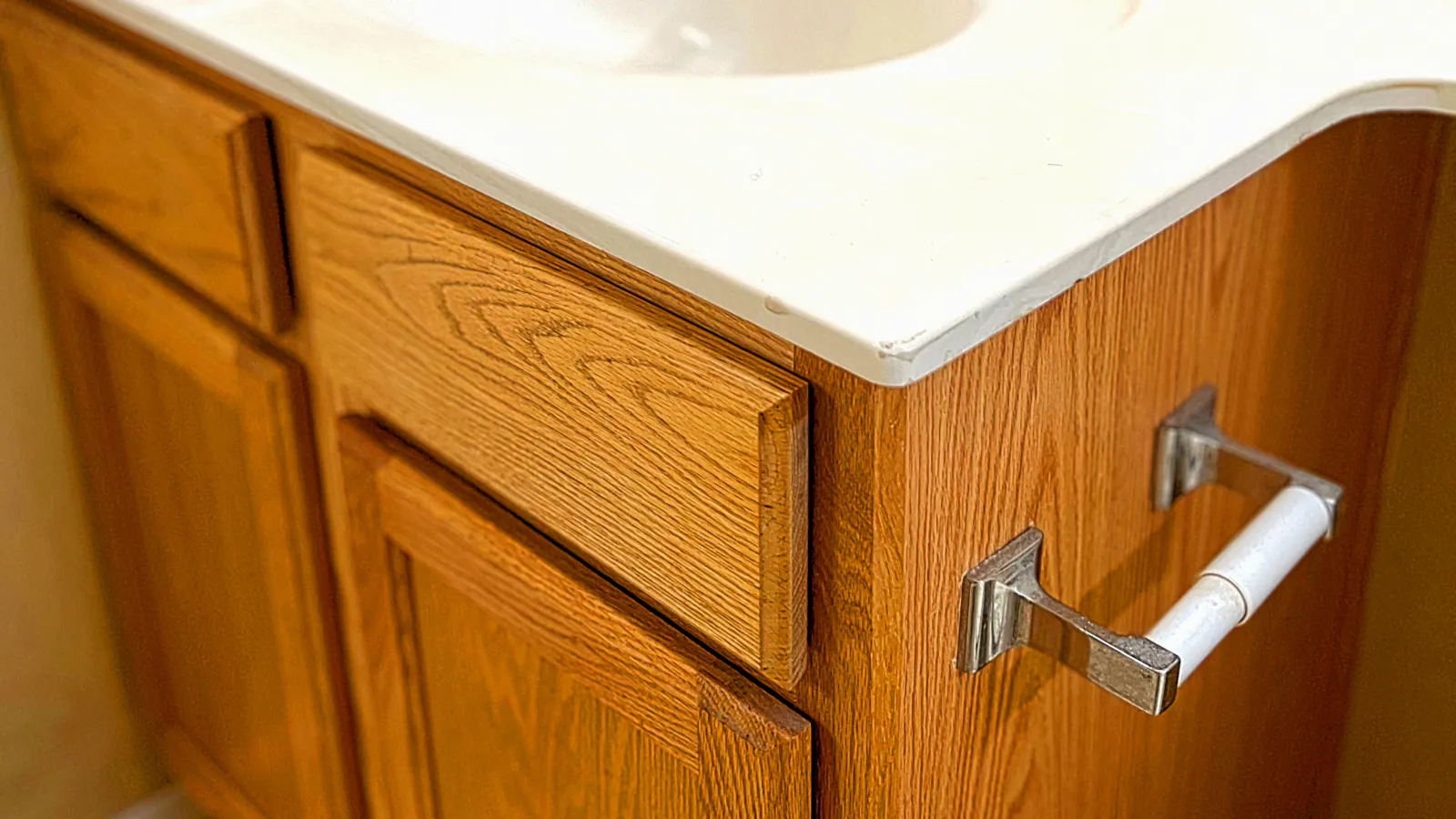 Bathroom sink with white countertop, silver faucet, wooden cabinet, and empty toilet paper holder.