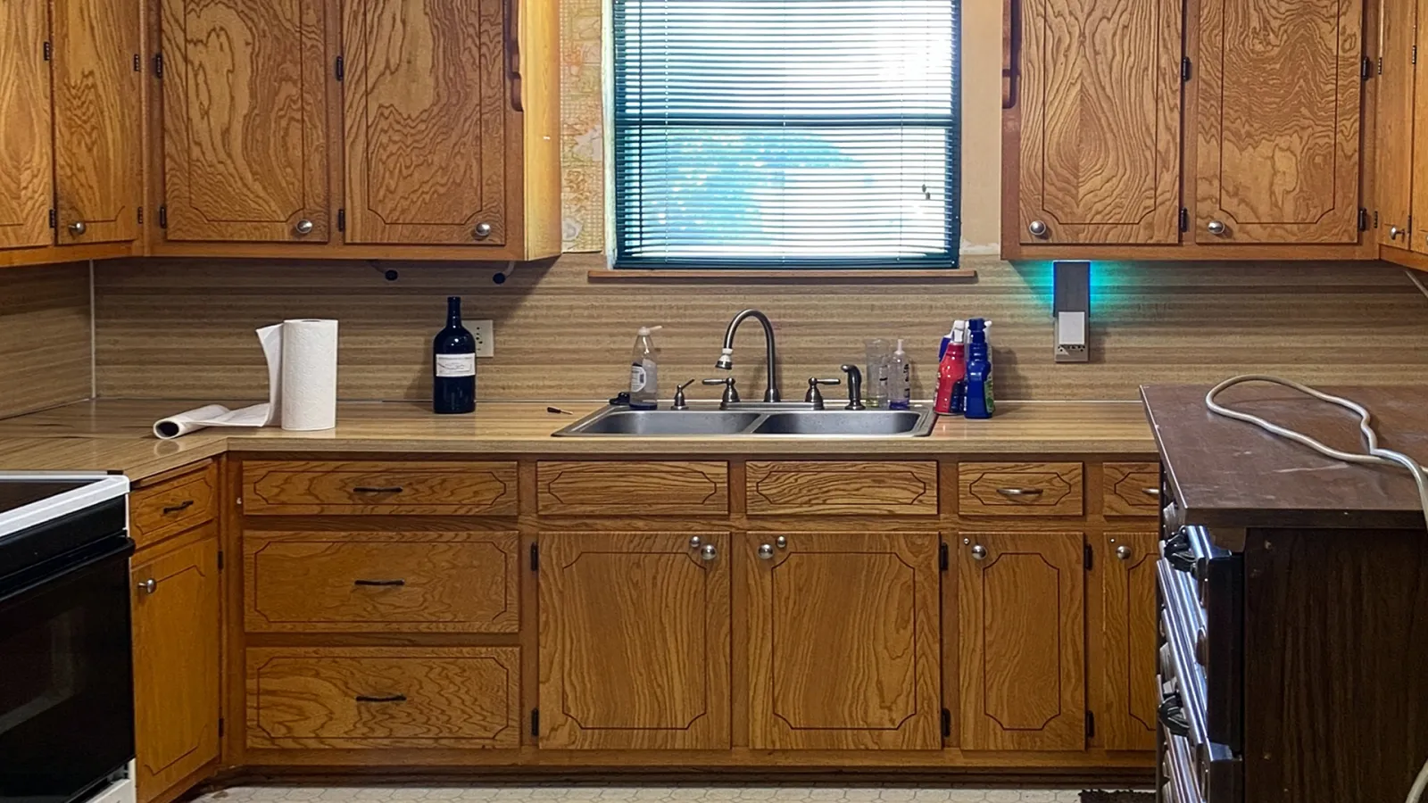 Vintage wooden kitchen cabinets with a double sink under window and various items on countertop including soap and paper towels.