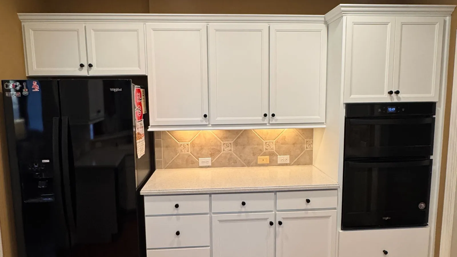 Modern kitchen section with white cabinets, black Whirlpool refrigerator and built-in double oven on hardwood floor.