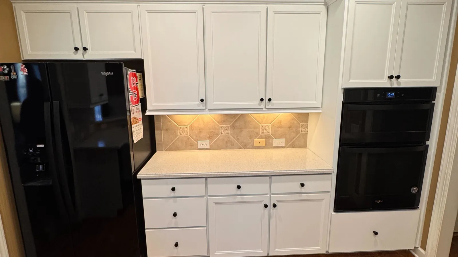 Modern kitchen with white cabinets, black appliances, wooden floors, and under-cabinet lighting over a tile backsplash.