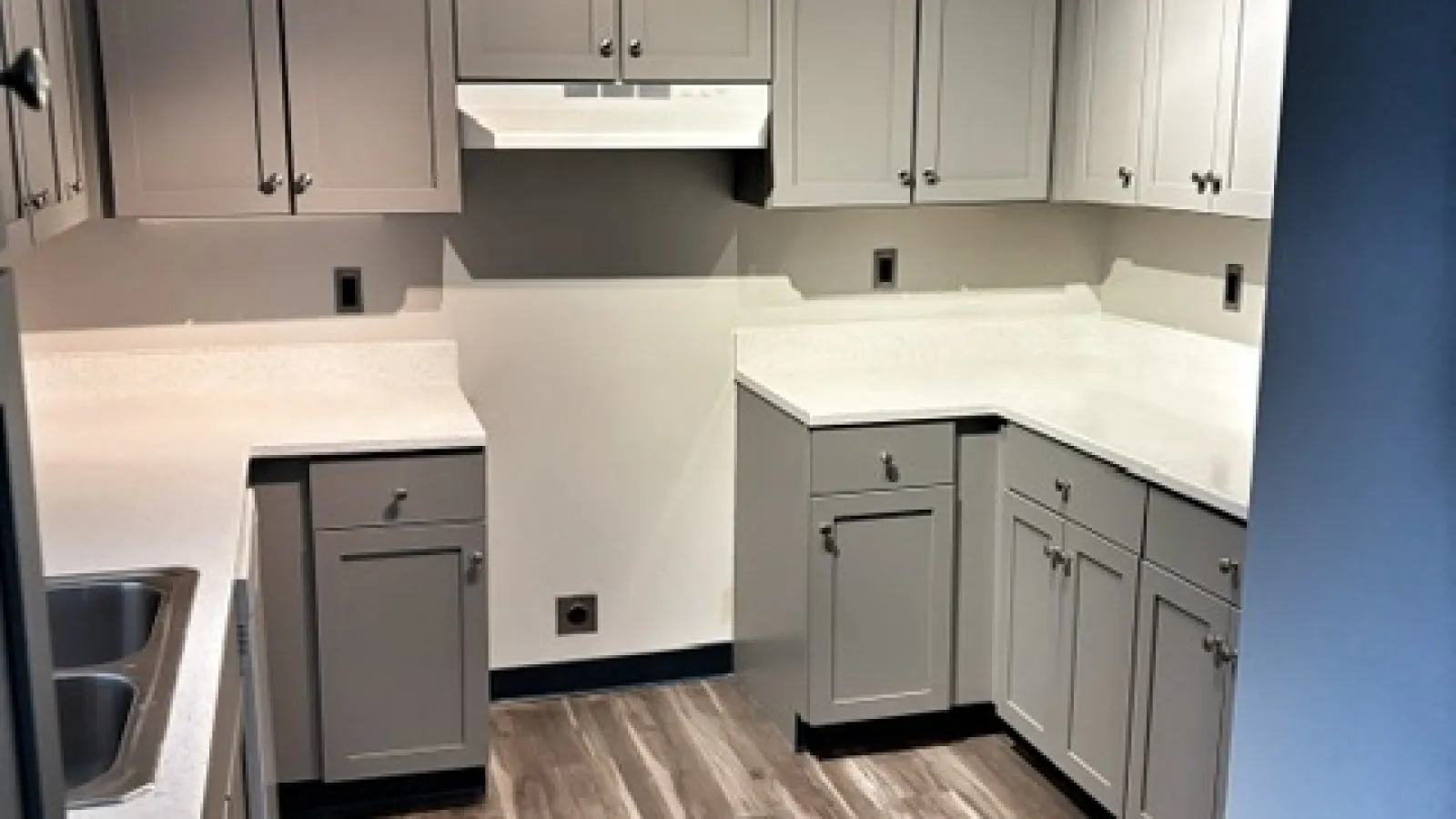 Modern kitchen with light gray cabinets, white countertops, stainless steel sink, and wood-look flooring under spotlight.
