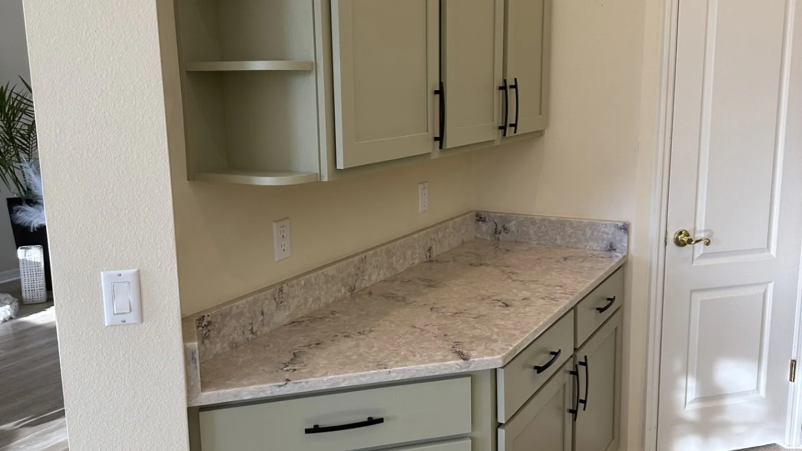 Kitchen corner with sage green cabinets, marble countertop, black handles, light wood flooring, and white door.