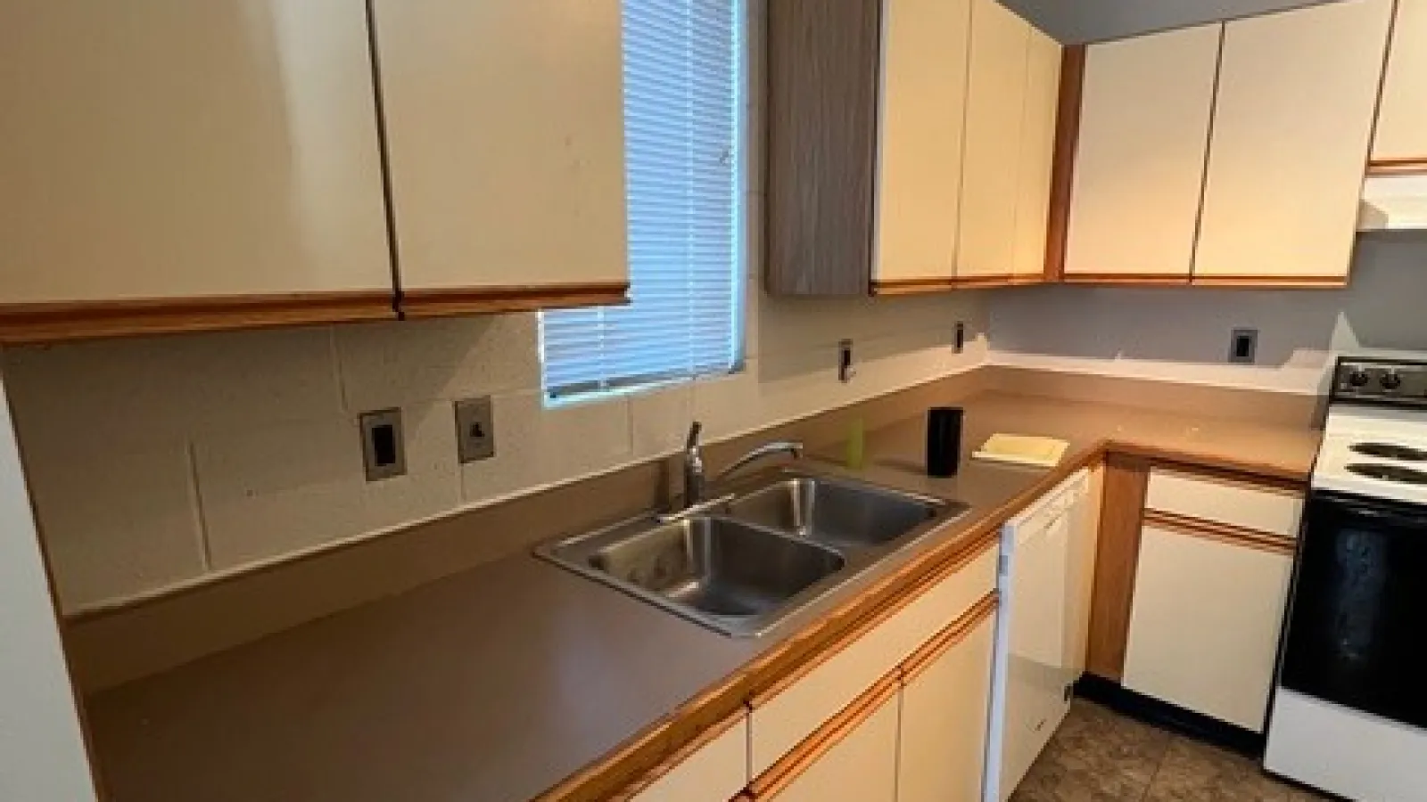 Small kitchen with beige cabinets, double sink, stove, refrigerator, and tiled floor with window blinds.