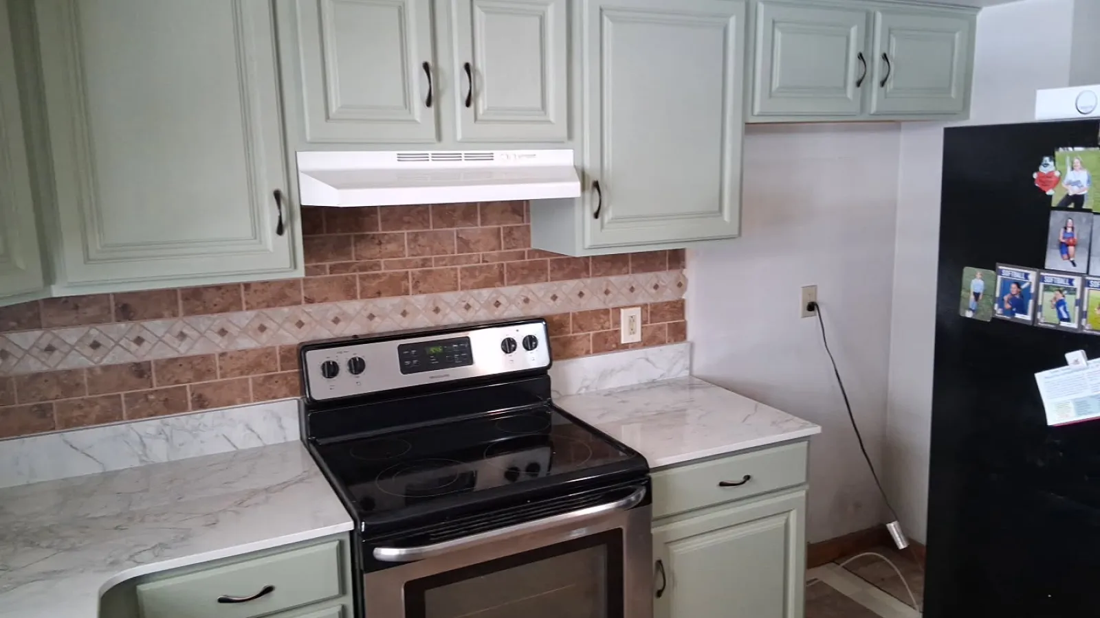 Modern kitchen with stainless steel stove, white marble countertops, sage green cabinets, and brown tiled backsplash.