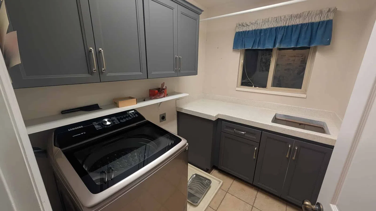 Laundry room with stainless steel washer, gray cabinets, white countertop, beige tiled floor, and window with blue valance
