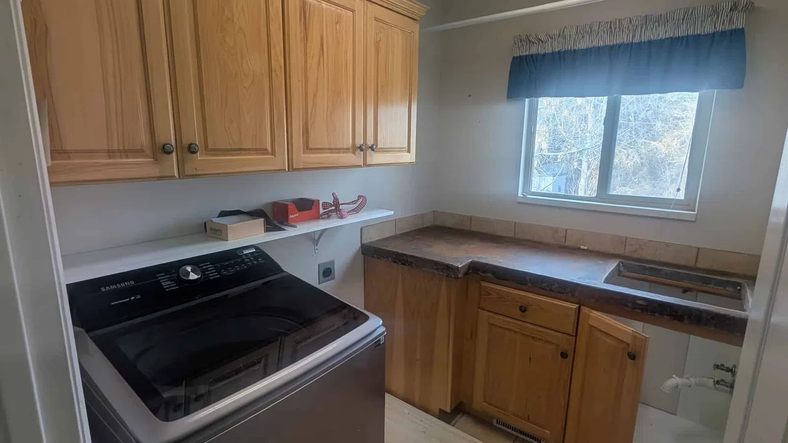 Laundry room with a Samsung washing machine, wooden cabinets, tiled floor, and a window with a blue valance.
