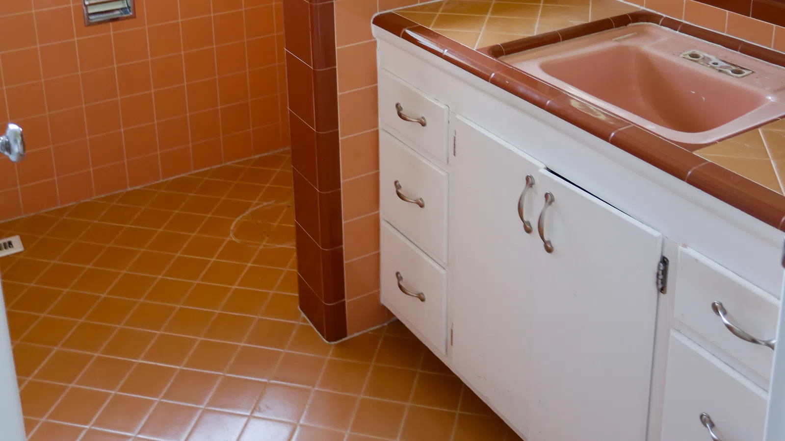 Retro bathroom with orange tile flooring and walls, pink sink, and white vanity with mirror and window.