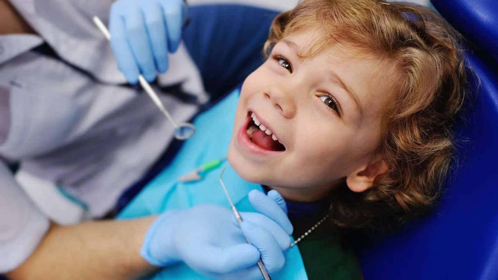 Smiling child with open mouth receiving dental checkup from dentist wearing blue gloves.