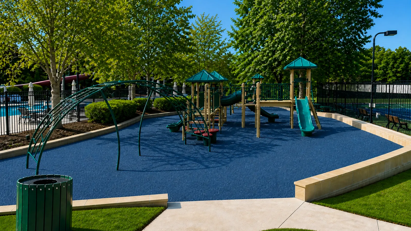 Outdoor playground with blue rubber flooring, green slides, climbing structures, and surrounding trees on a sunny day