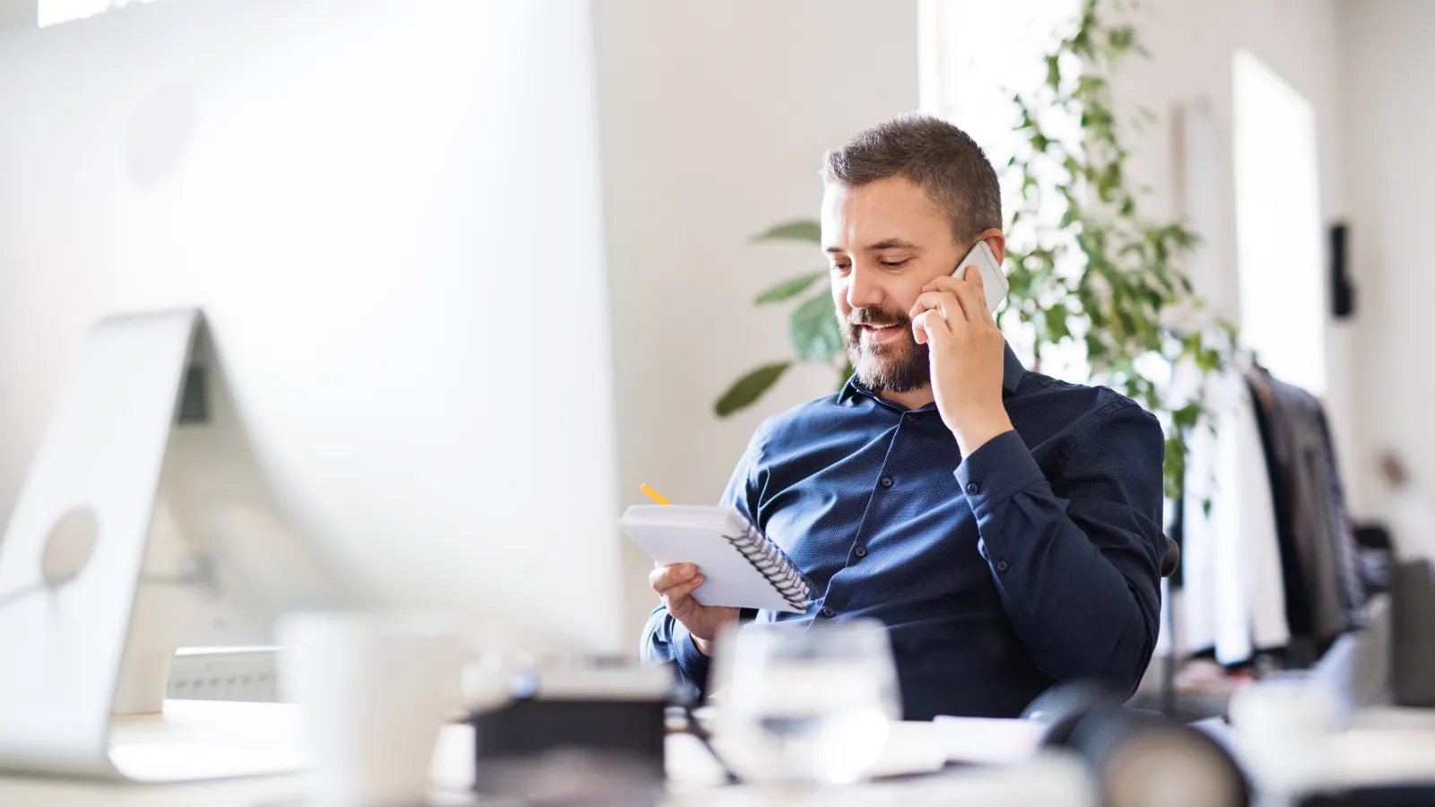 Man in blue shirt talking on phone and taking notes in a bright modern office with plants.