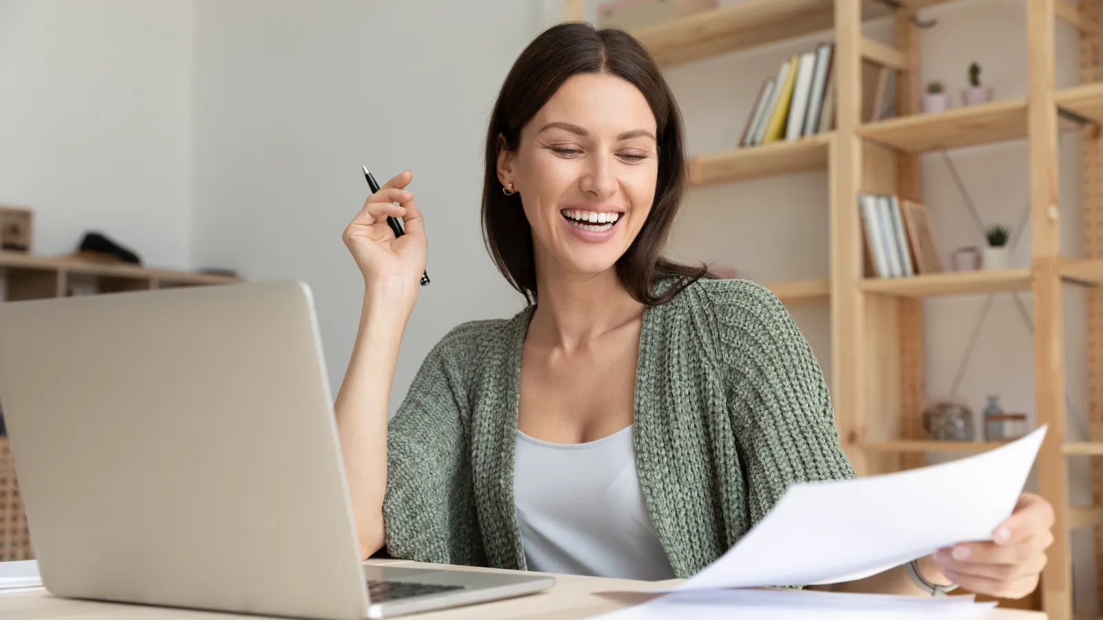 Happy woman working at home office with laptop and paperwork, smiling and holding a pen