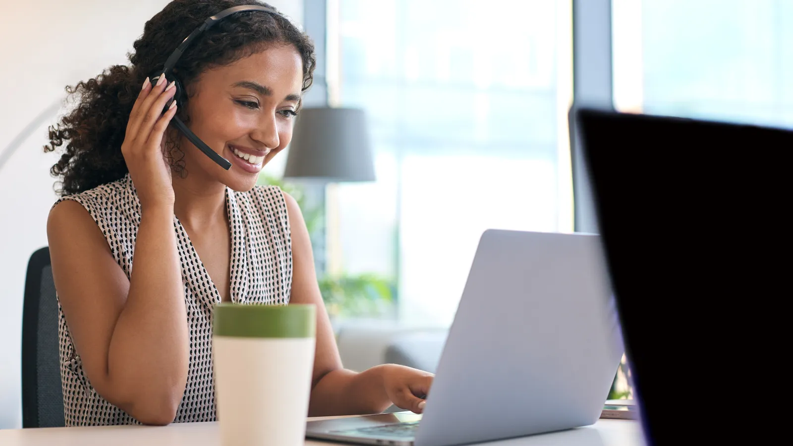 Smiling woman wearing headset working on laptop at bright office desk with coffee cup nearby