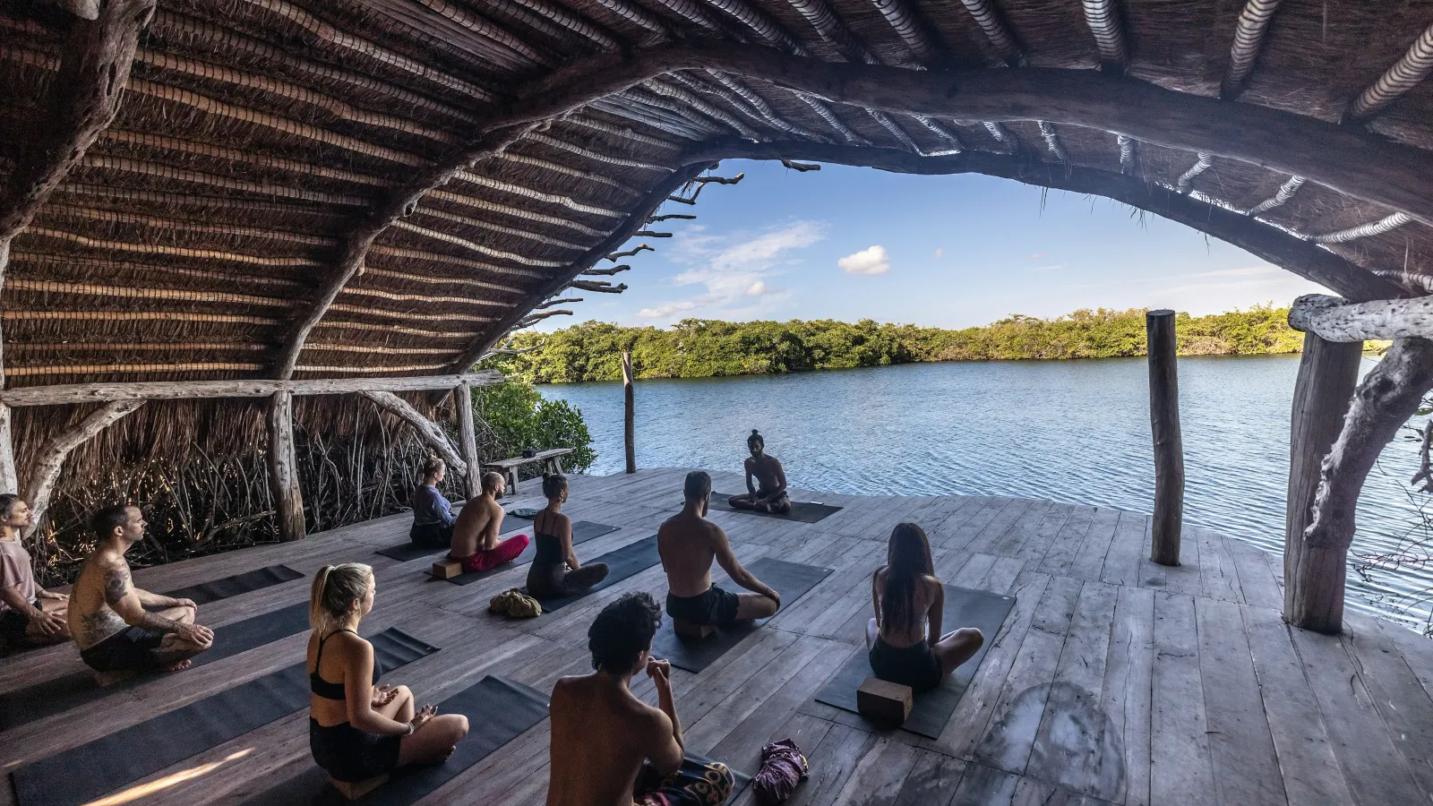 Group meditation session under a thatched roof by a lake with nature in the background at sunset.