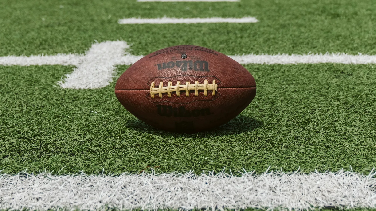 Wilson football resting on green turf field near white yard lines during daytime game.