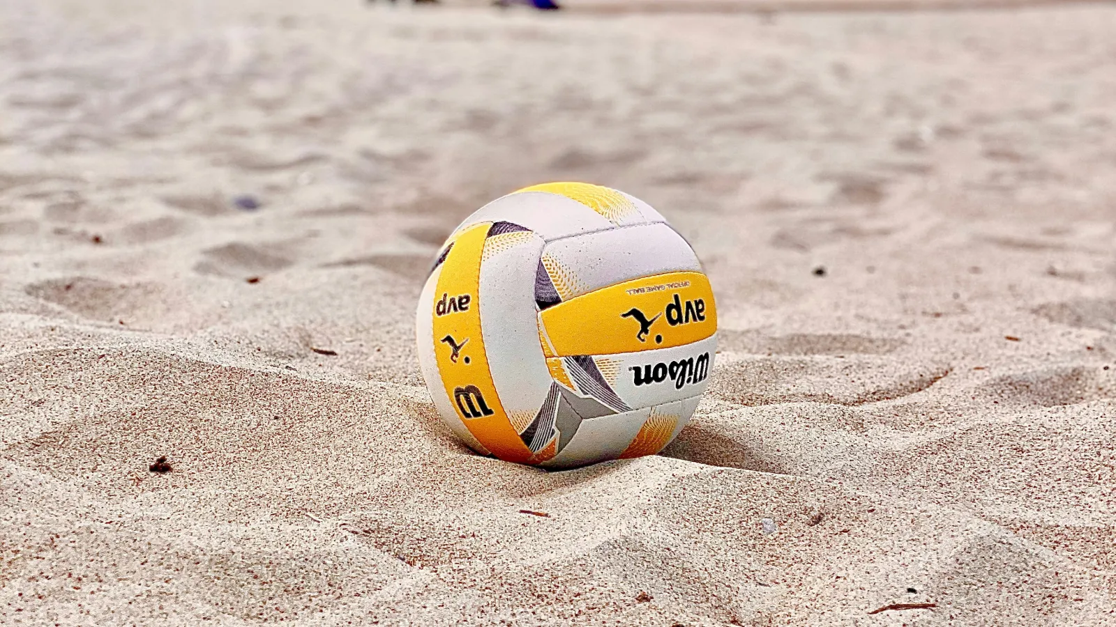Yellow and white volleyball resting on sandy beach with dunes and houses in background under cloudy sky.