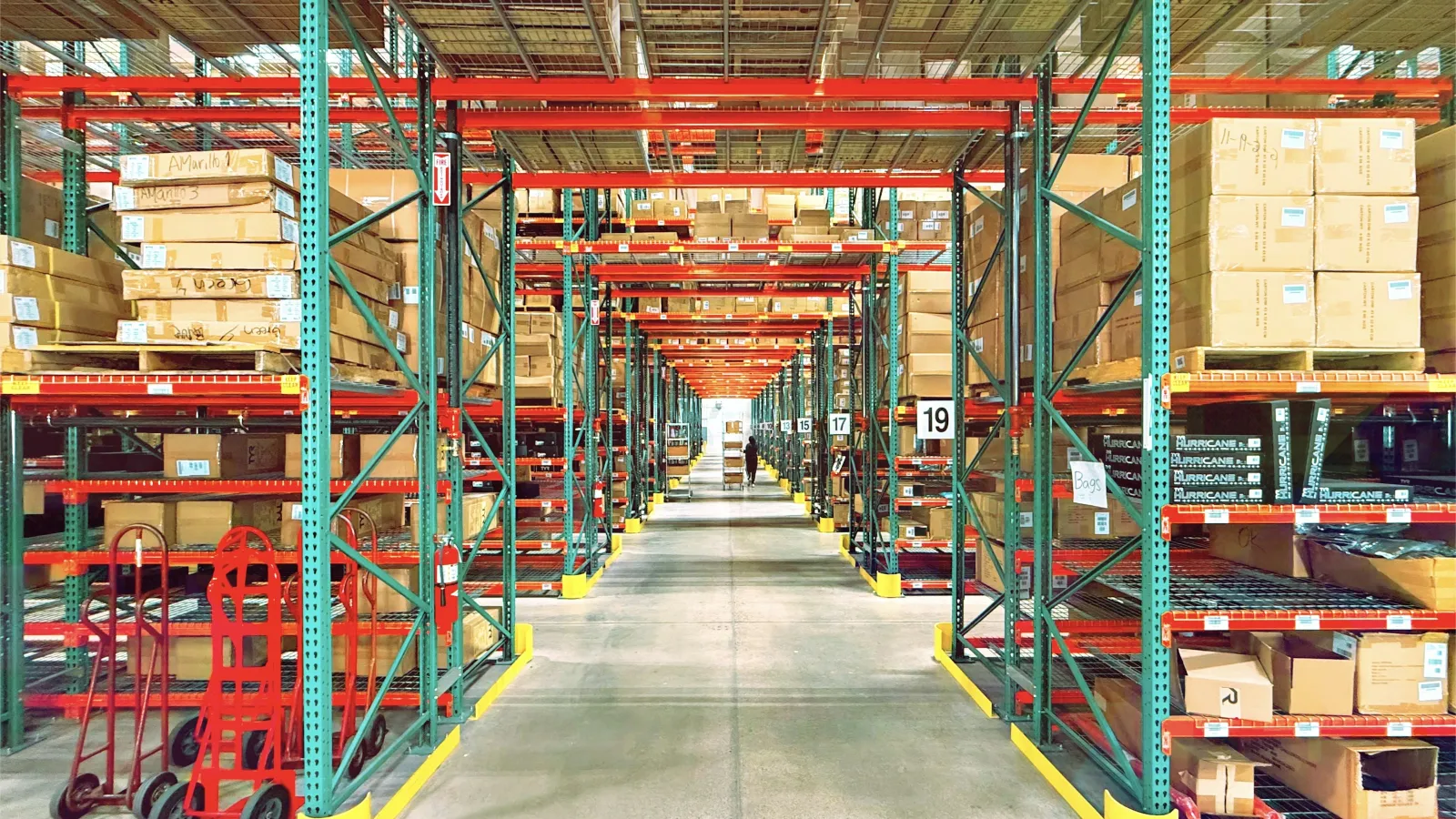 Spacious warehouse aisle with shelves stocked with boxes and hand trucks lined up on the side under bright lighting.
