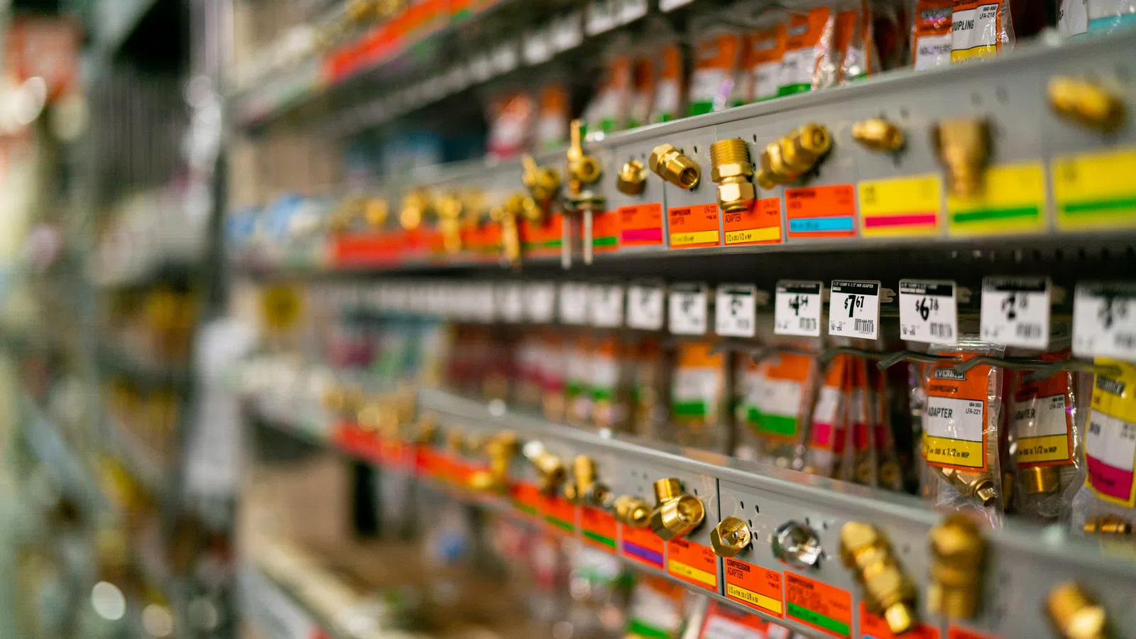 Shelves filled with brass plumbing fittings and parts with colorful price tags in a hardware store aisle.
