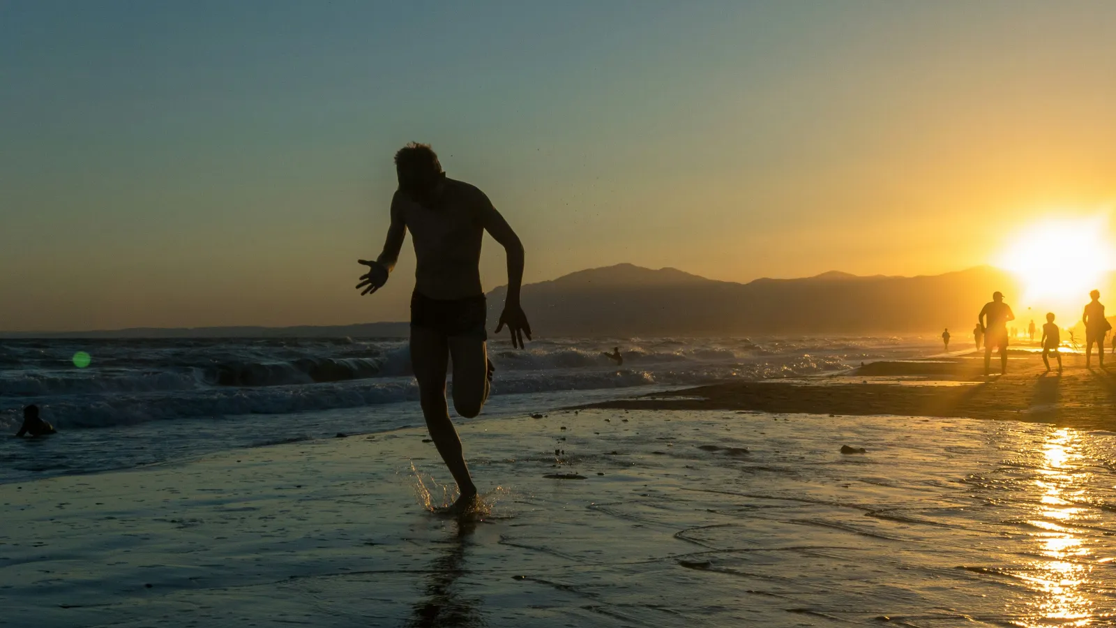 Silhouetted man running on wet beach at sunset with waves and people in background.