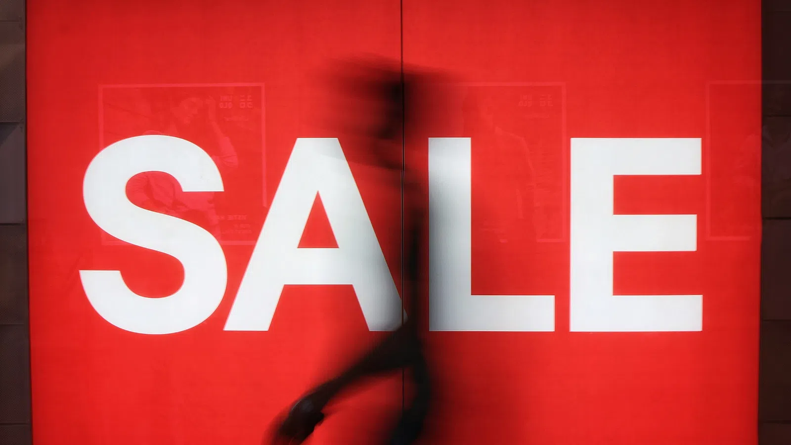 Blurred silhouette walking past large white SALE sign on bright red background in store window.