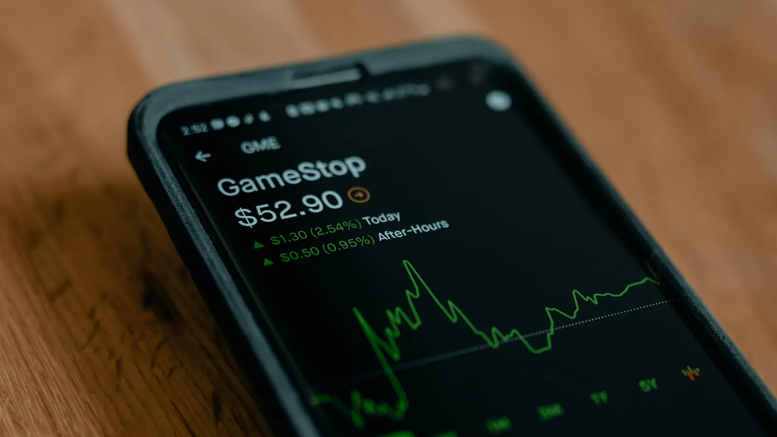 Smartphone screen showing GameStop stock price at $52.90 with green upward trend graph on wooden surface.