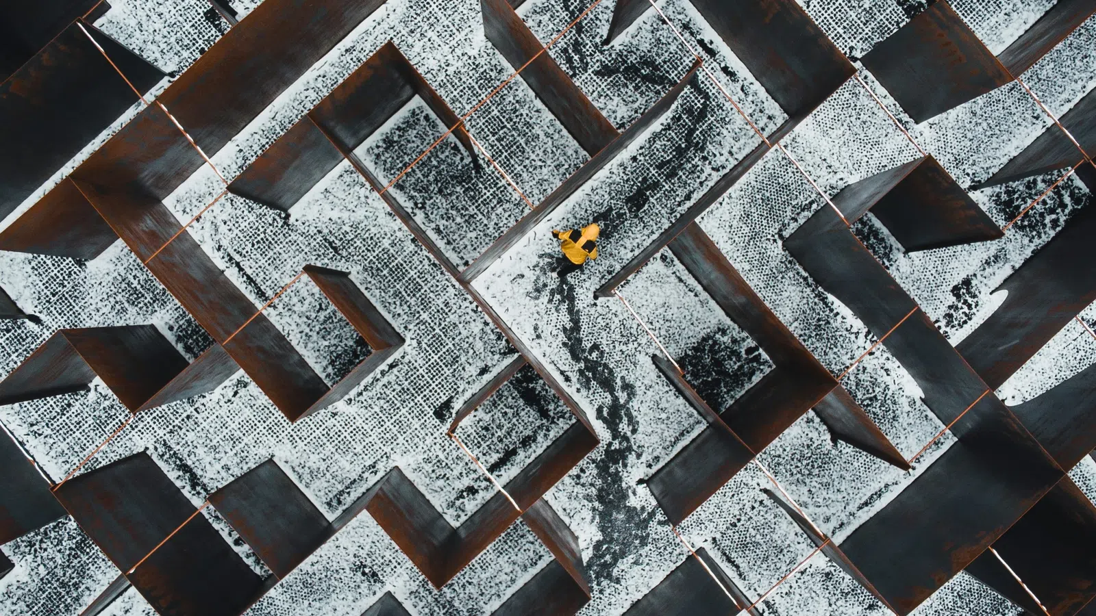 Aerial view of a person in a yellow jacket navigating through a snow-dusted metal maze with rusted walls.