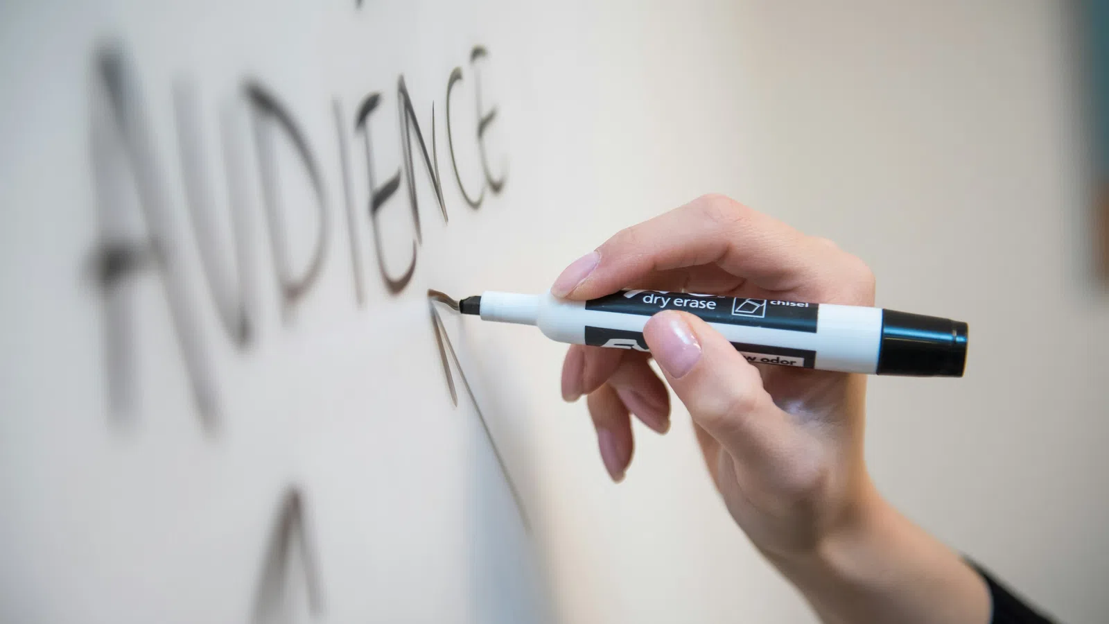 Hand holding a black dry erase marker writing the word AUDIENCE on a whiteboard.