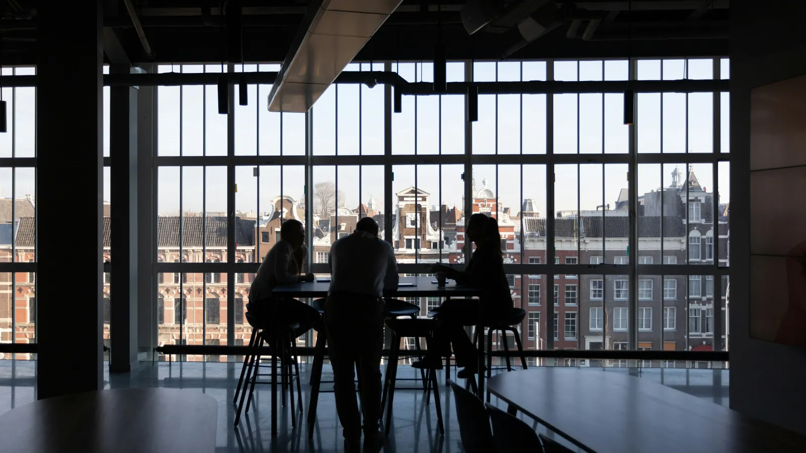 Silhouettes of three people sitting and standing around a table in a modern office with large windows and city views.
